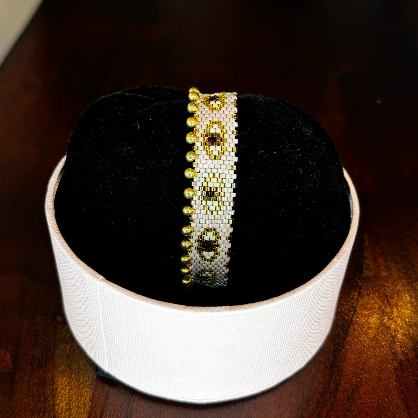 A bracelet displayed on a person's wrist and on a flat surface, featuring a nude color with gold accents and small white beads.