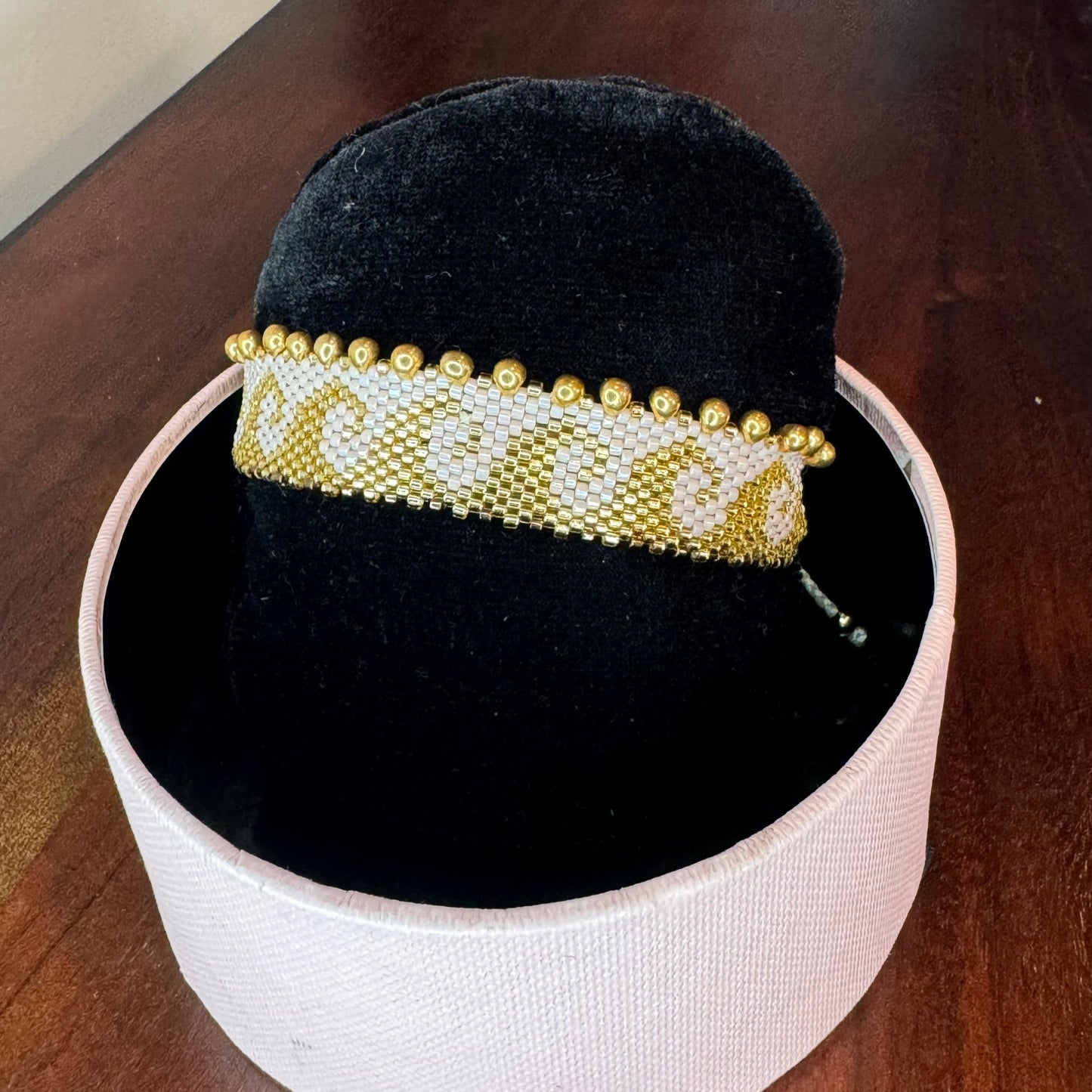 A bracelet displayed on a person's wrist and on a flat surface, featuring a nude color with gold accents and miyuki rose and gold beads.