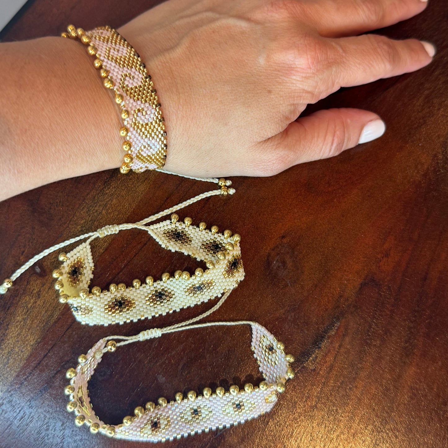 A bracelet displayed on a person's wrist and on a flat surface, featuring a nude color with gold accents and small white beads.