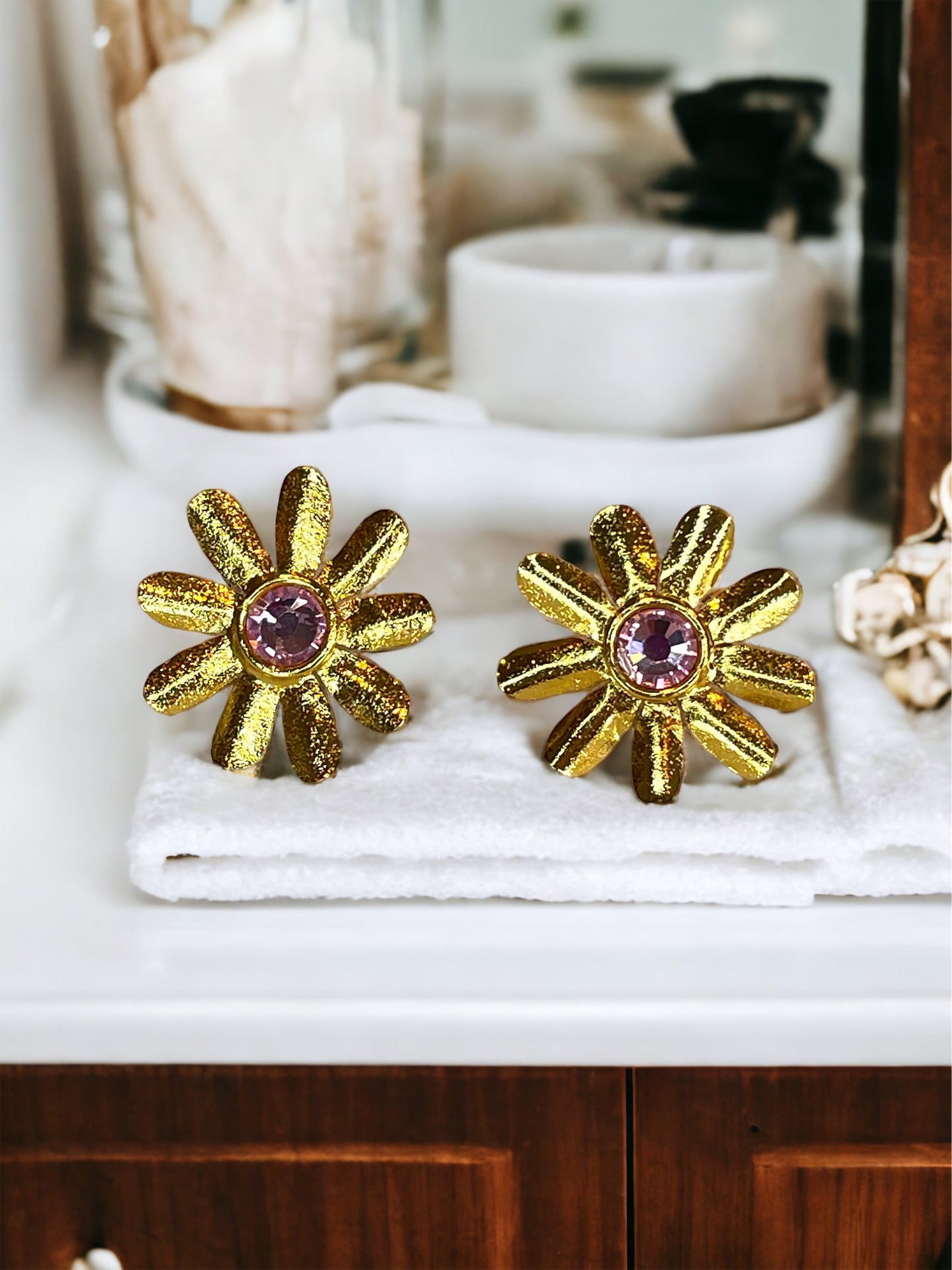 A pair of gold-plated bronze daisy-shaped earrings with a light pink Swarovski crystal in the centre, displayed on a bathroom counter.