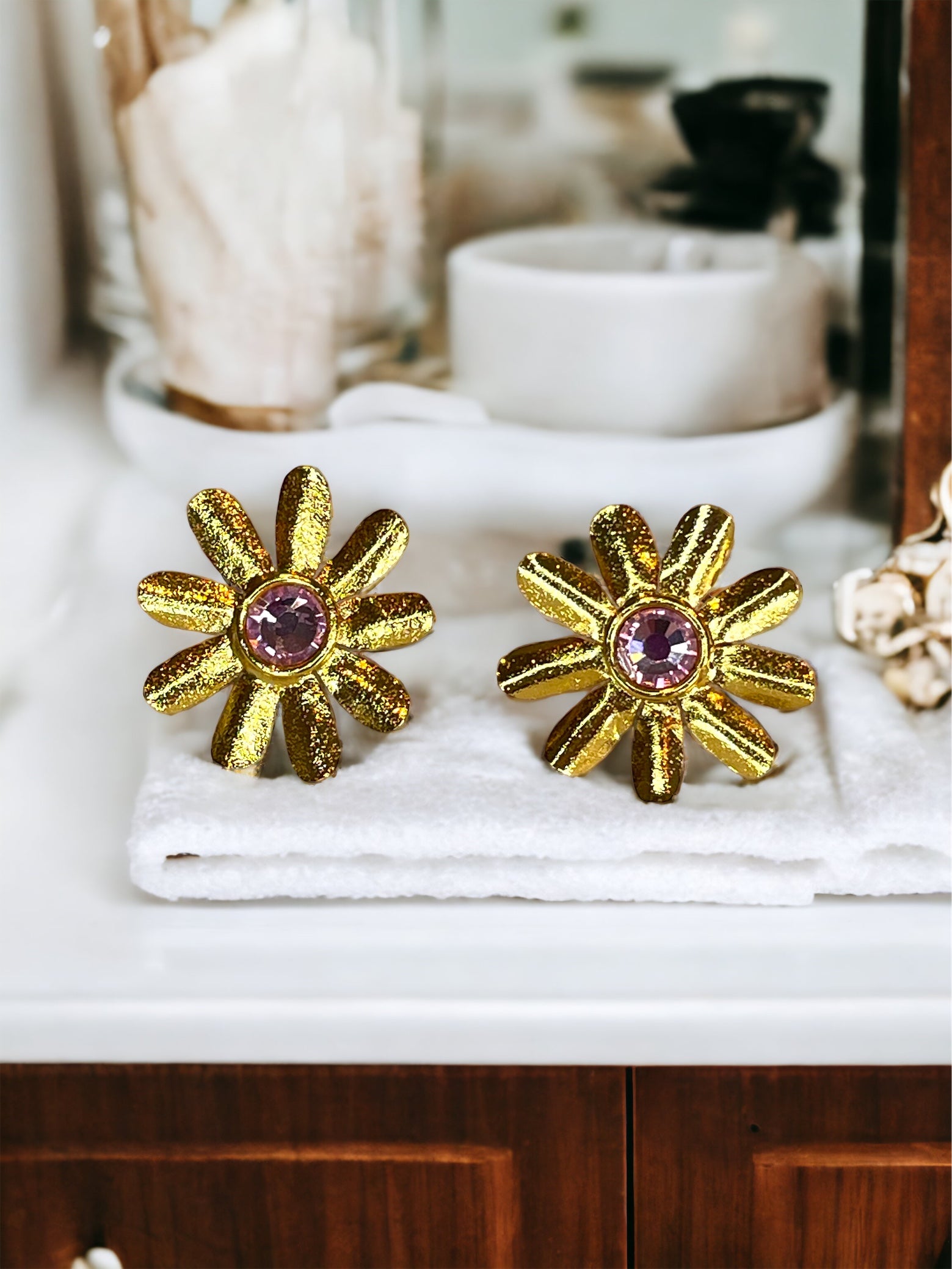 A pair of gold-plated bronze daisy-shaped earrings with a light pink Swarovski crystal in the centre, displayed on a bathroom counter.