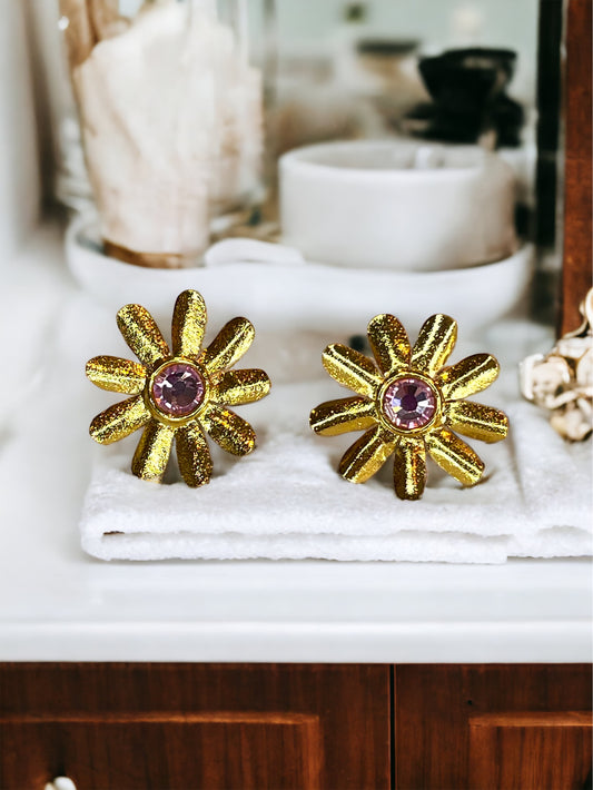 A pair of gold-plated bronze daisy-shaped earrings with a light pink Swarovski crystal in the centre, displayed on a bathroom counter.