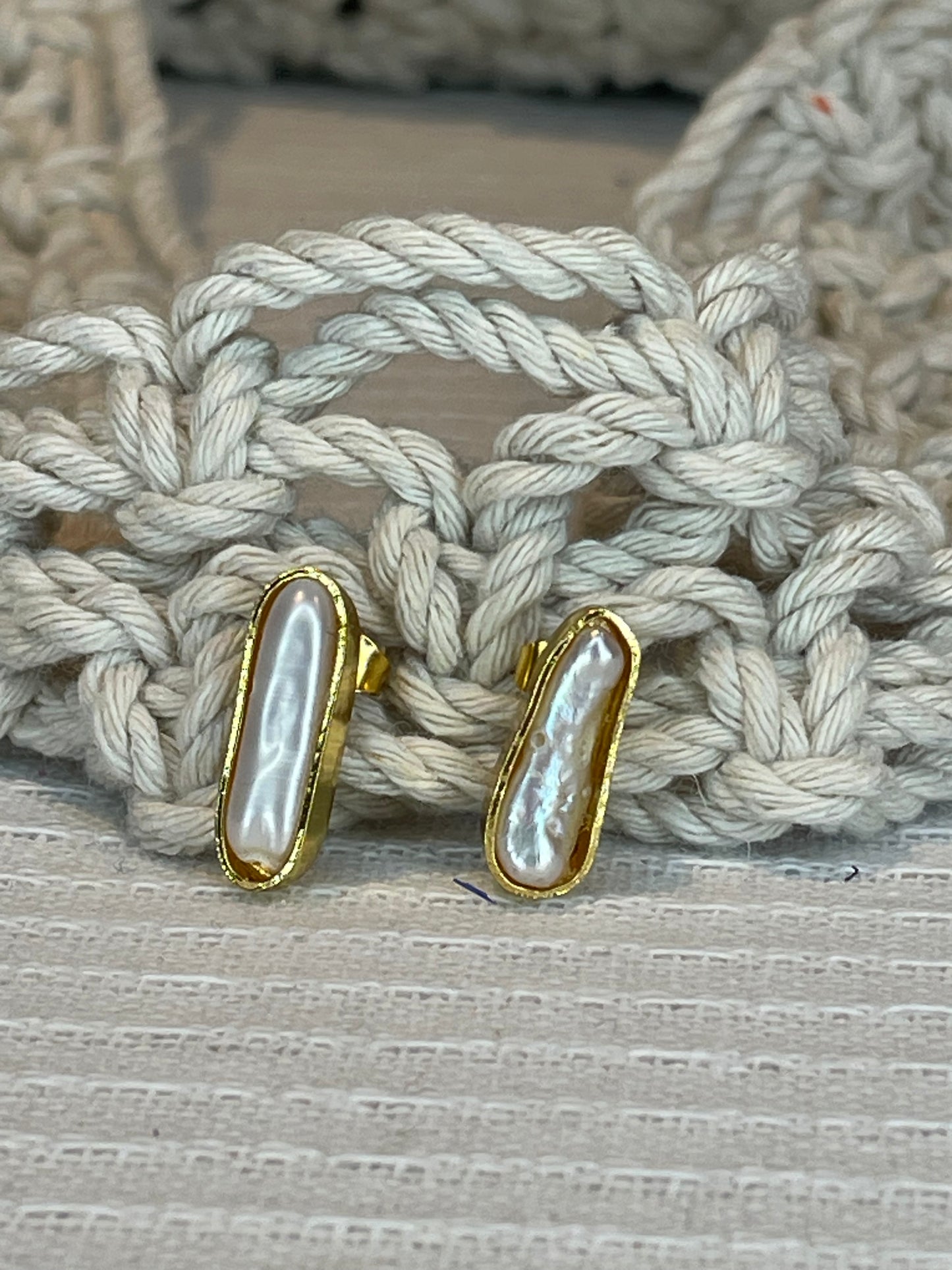 A pair of pearl stick studs with gold plating and rope-like texture on the back, displayed on a textured surface.