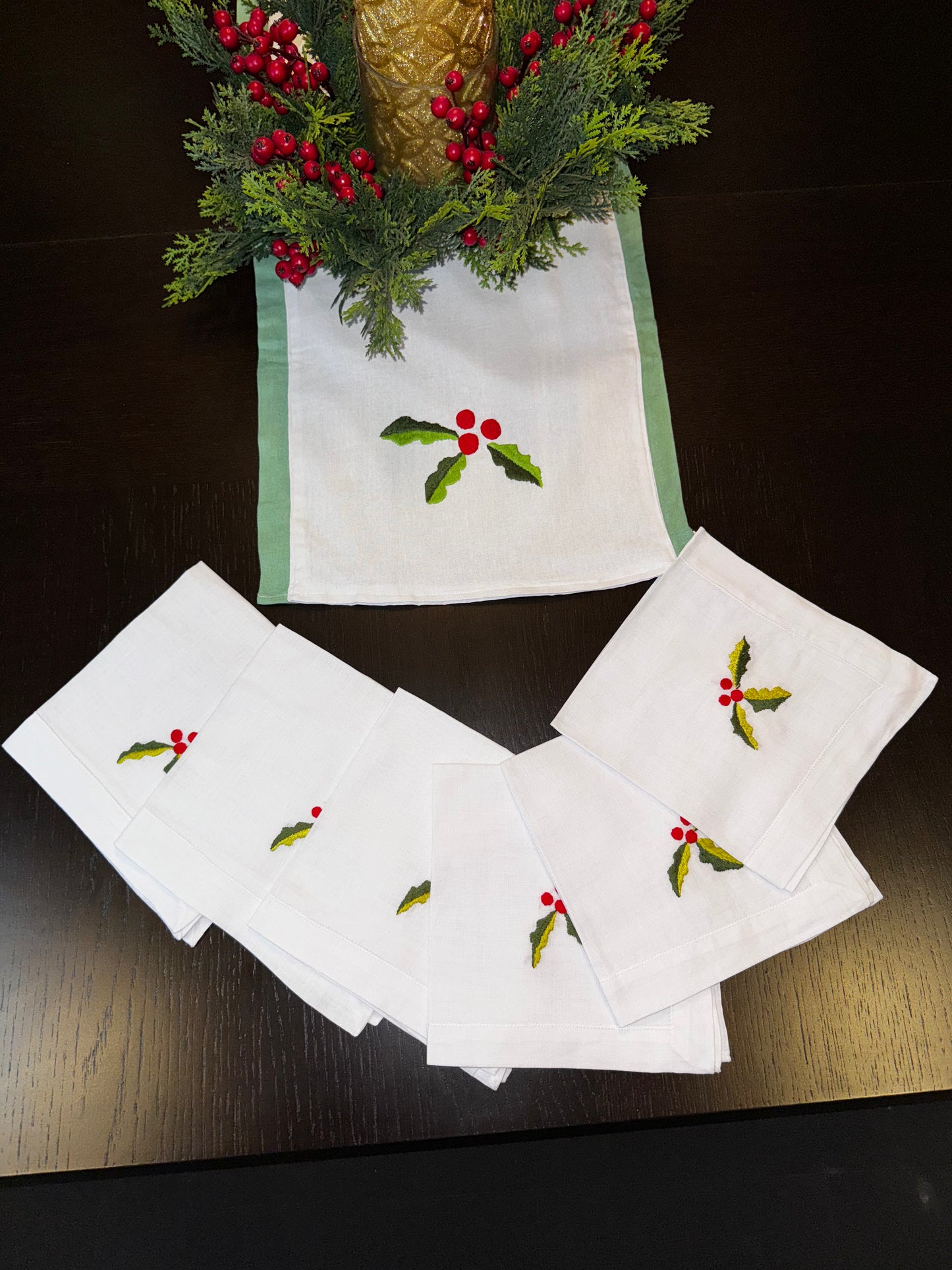 A set of white linen napkins with red Christmas-themed embroidery, displayed on a table with a festive table runner and decorative branches in the background.