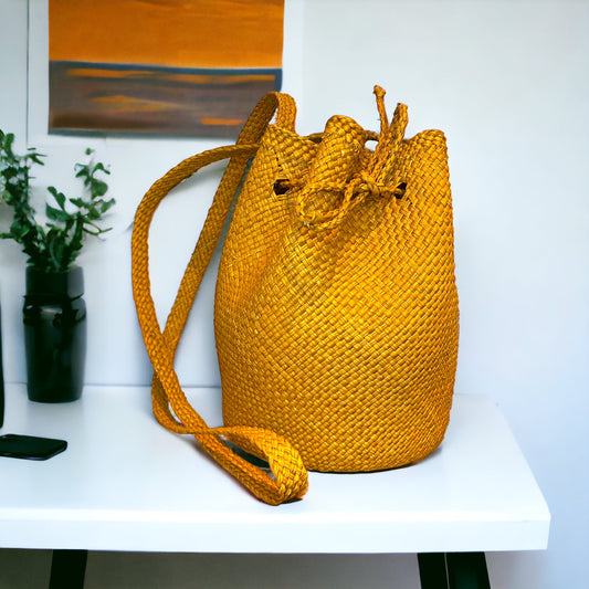 A yellow wicker bucket bag with a drawstring closure, placed on a white surface with a blurred background.