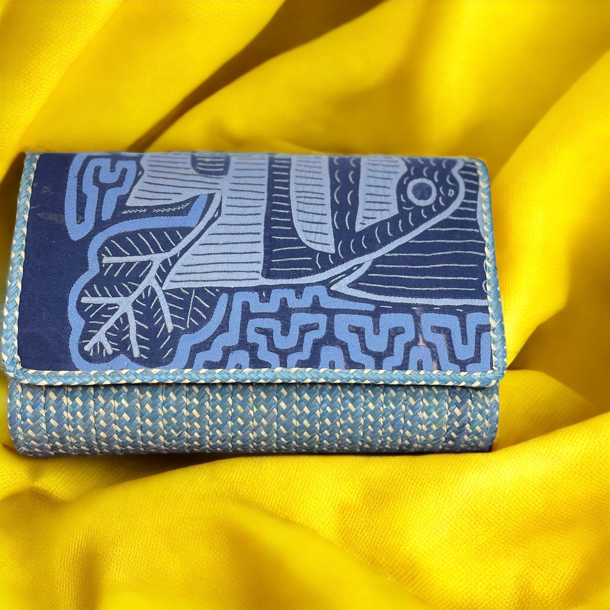 A blue and yellow clutch with a geometric mola pattern and a strap, placed on a yellow fabric surface.