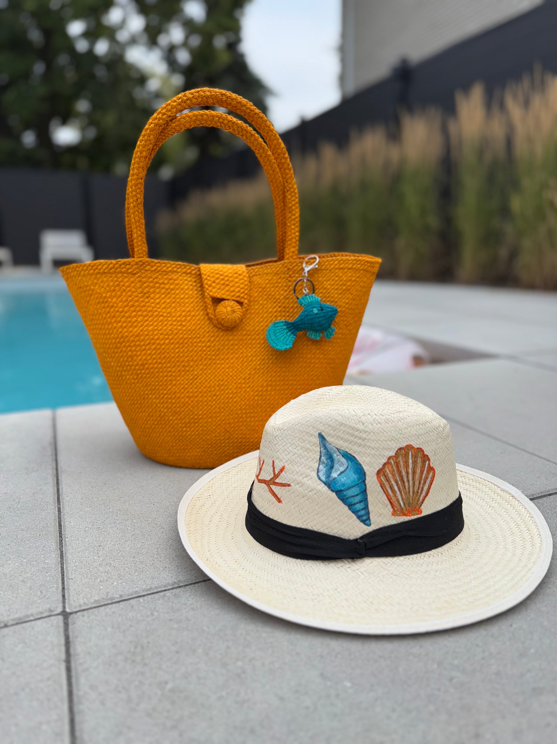 dark yellow woven bag with a blue keychain and a white hat with shell designs on a poolside surface.