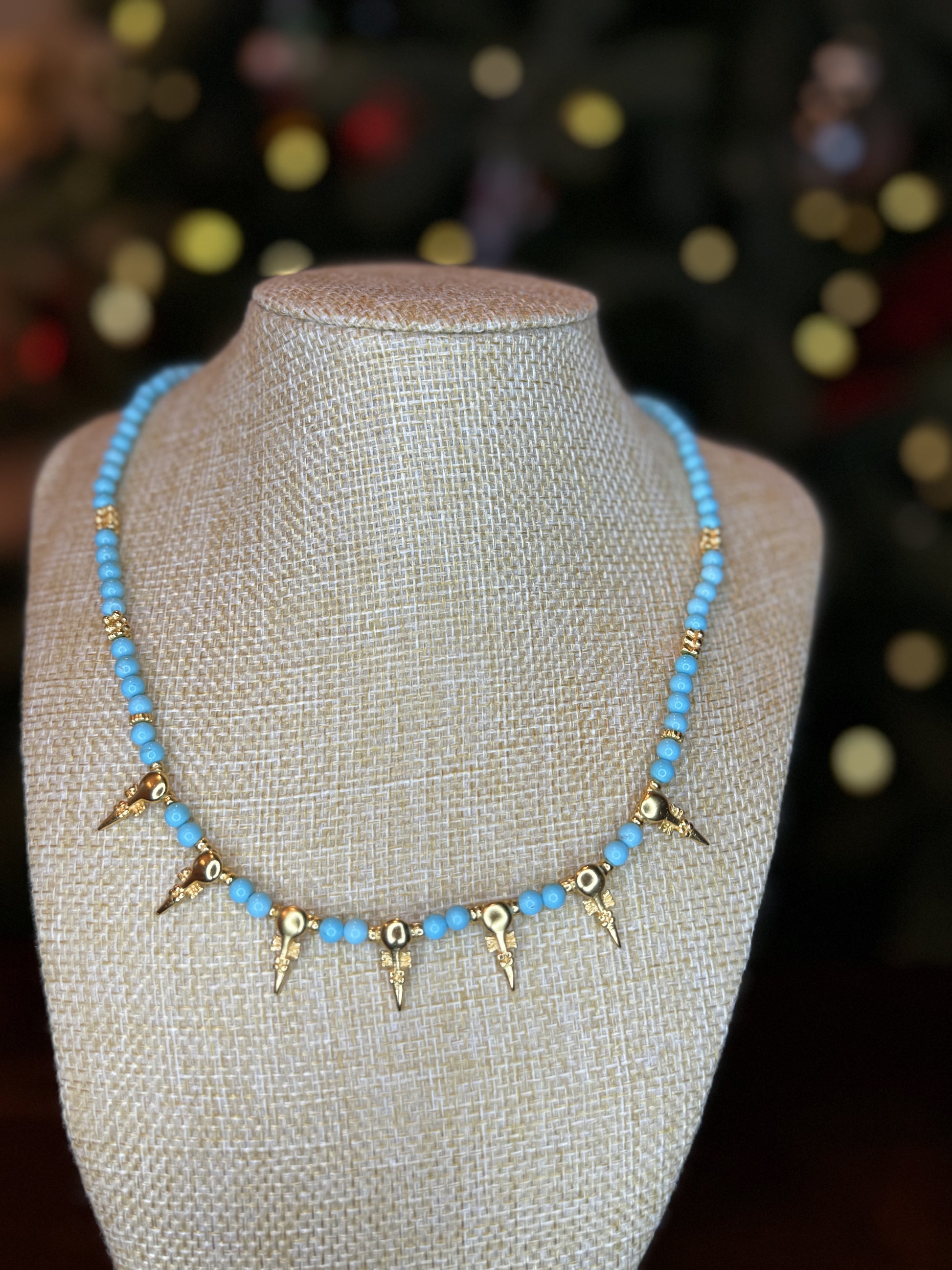 A turquoise beaded necklace with gold-plated accents displayed on a mannequin. The necklace features a unique design with geometric shaped pendants.