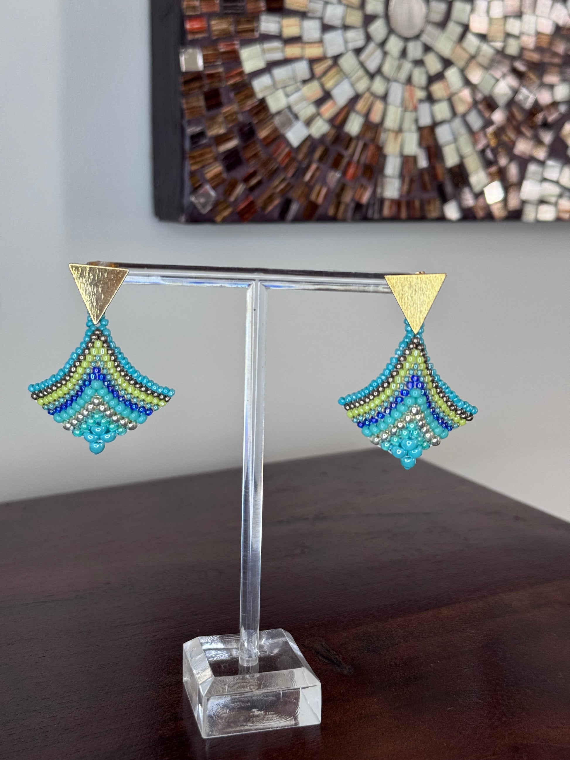 A pair of triangular-shaped aquamarine, gold, and green beaded earrings on display against a tiled background.