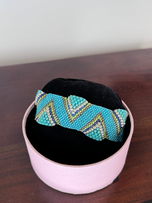 A beaded bracelet with aquamarine, gold, and green hues, displayed on a black cushion inside a pink box.