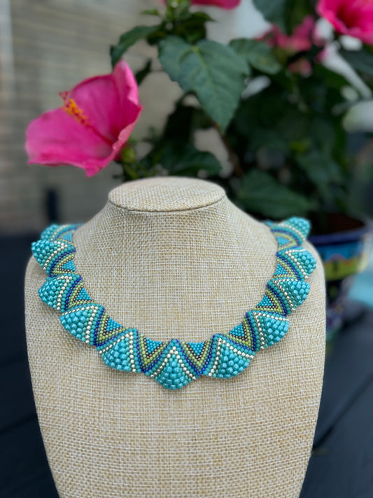 beaded necklace with aquamarine, gold and green hues, displayed on a mannequin outdoors. The necklace has a geometric pattern and a toggle clasp.