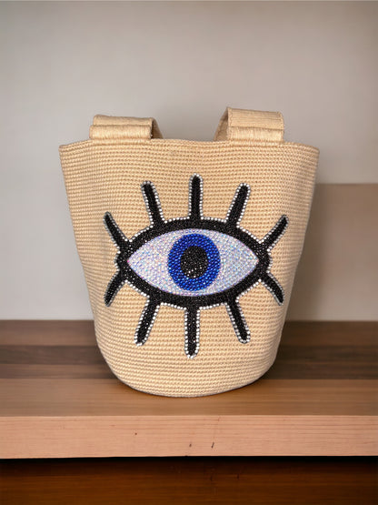 A beige-colored, flat-weave cotton bag featuring a decorative 'evil eye' design with crystals embedded in the pattern, set against a wooden background.