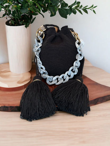 A black Lux Mochila handbag with a chain strap and tassel detail displayed on a wooden surface.