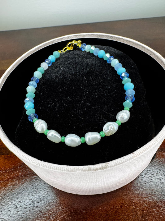 A beaded bracelet  featuring light blue and turquoise faceted crystal beads, white baroque pearls, small green beads, and a gold clasp.
