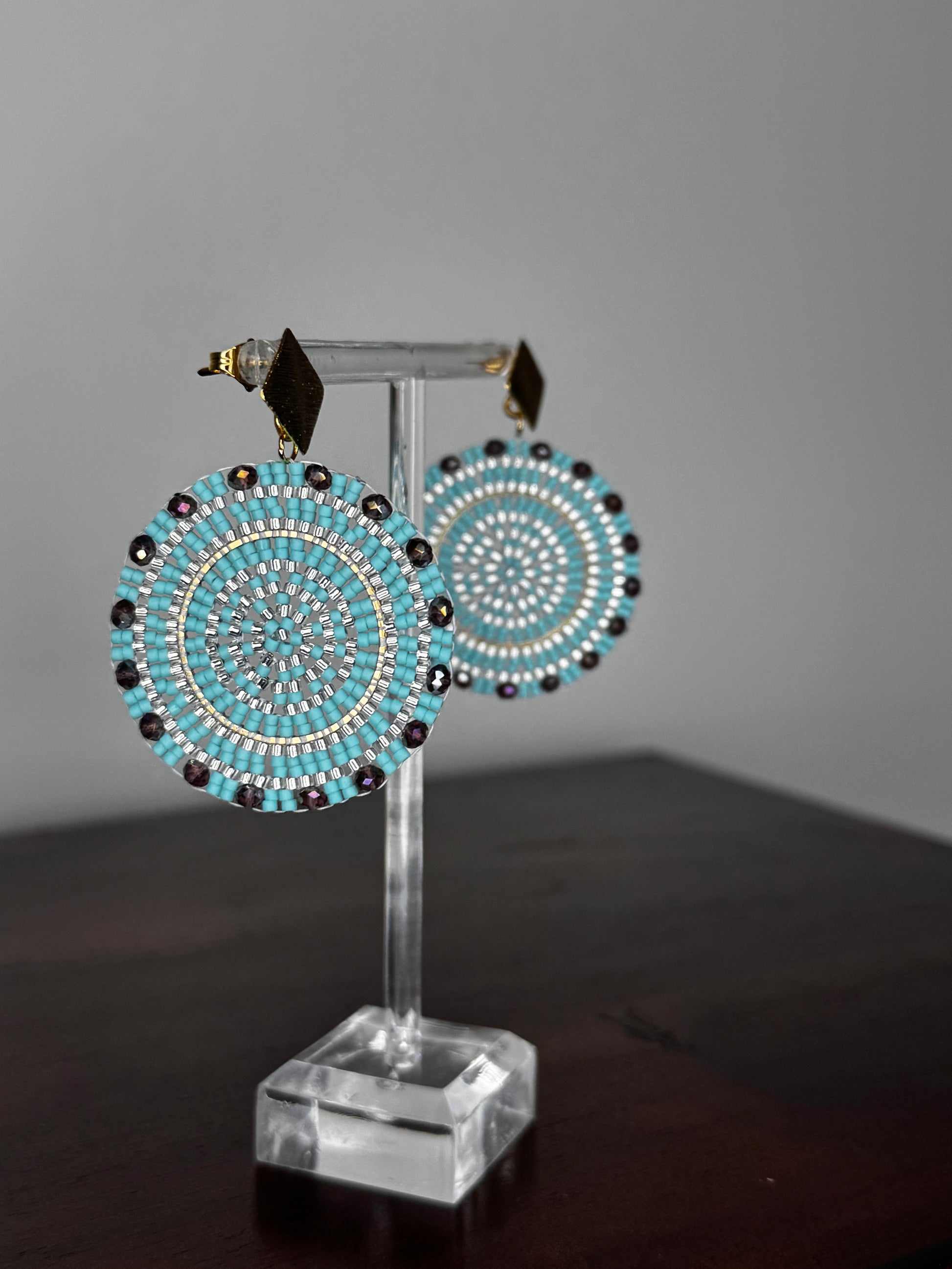 A pair of Spark Earrings displayed on a stand, featuring a hypoallergenic German silver stud and blue acrylic crystals in a circular beaded design.