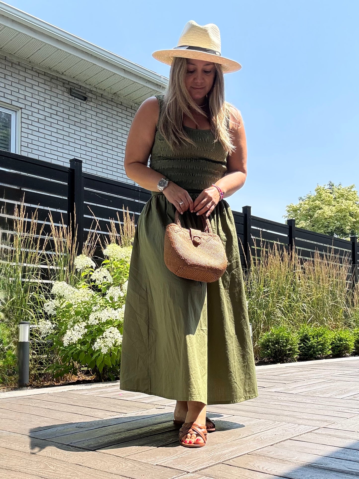 Woman in a green dress and hat holding a brown handbag on a wooden deck.