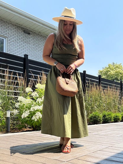 Woman in a green dress and hat holding a brown handbag on a wooden deck.