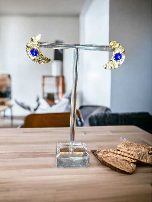 A pair of gold-plated bronze earrings with a central Swarovski blue crystal, displayed on a clear stand.