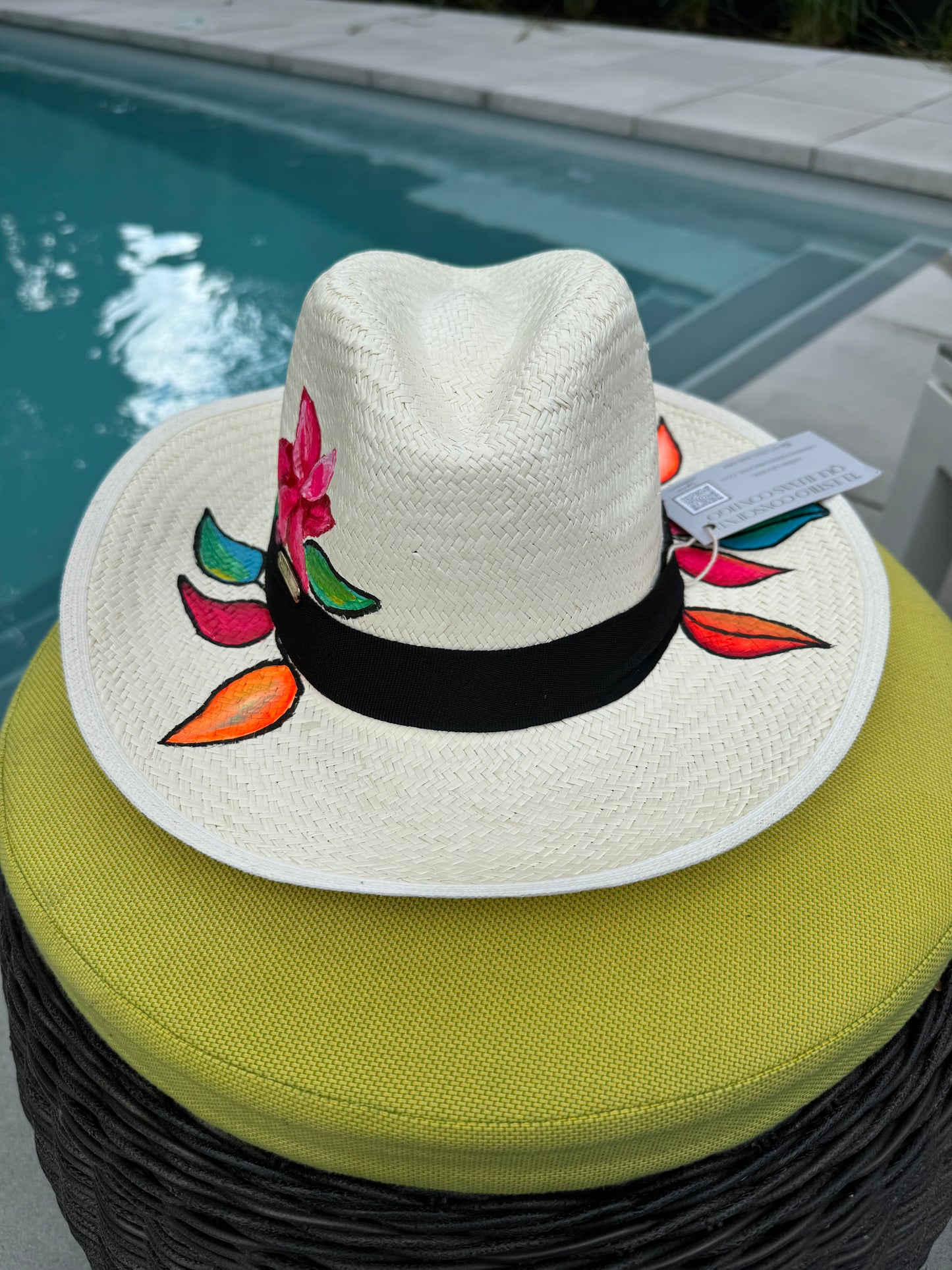 A hand-painted straw hat with a black band, featuring a floral design, resting on a wicker chair by a pool.