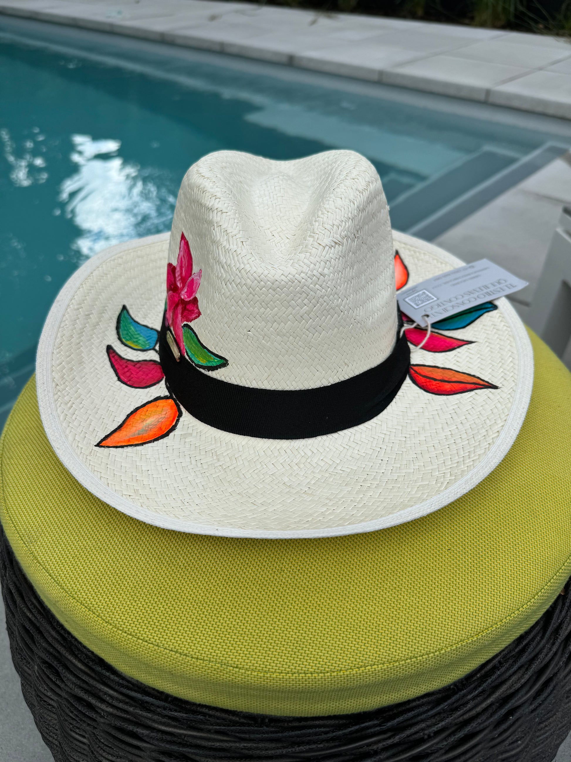 A hand-painted straw hat with a black band, featuring a floral design, resting on a wicker chair by a pool.