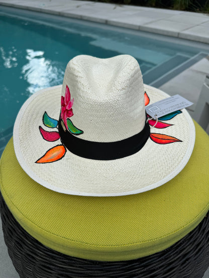 A hand-painted straw hat with a black band, featuring a floral design, resting on a wicker chair by a pool.