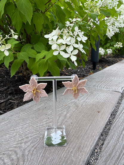 beige flower-shaped earrings on a clear stand with green foliage in the background