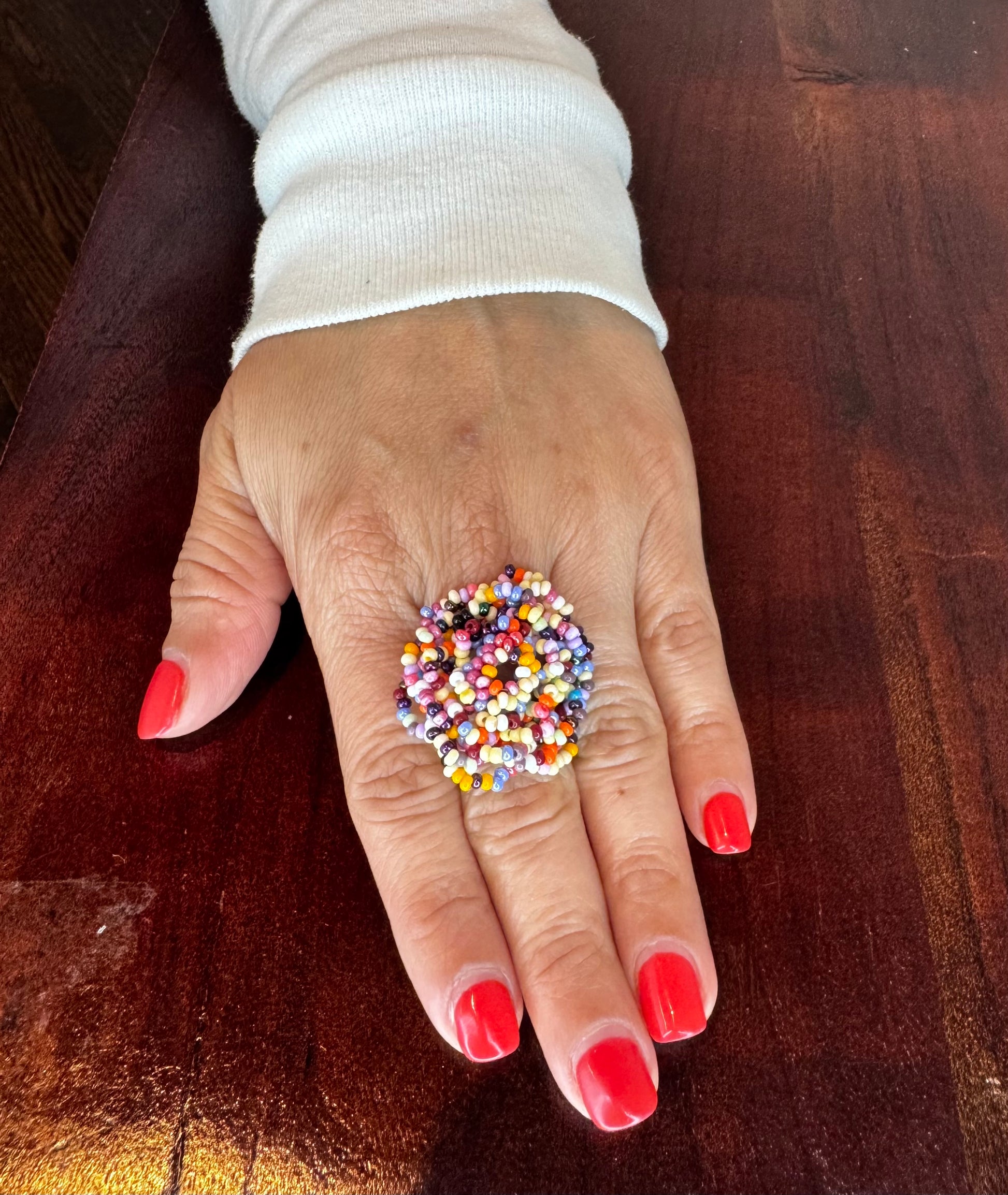 A colorful woven beaded ring displayed on a person's finger with red nail polish.