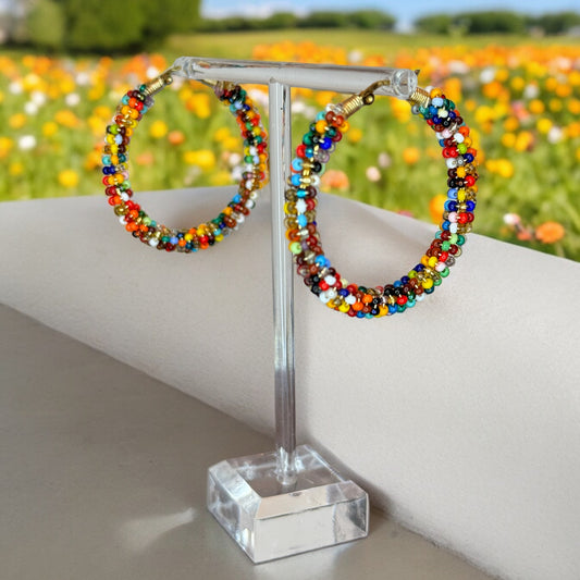 A pair of multicolored beaded hoop earrings displayed on a stand with a background of flowers.