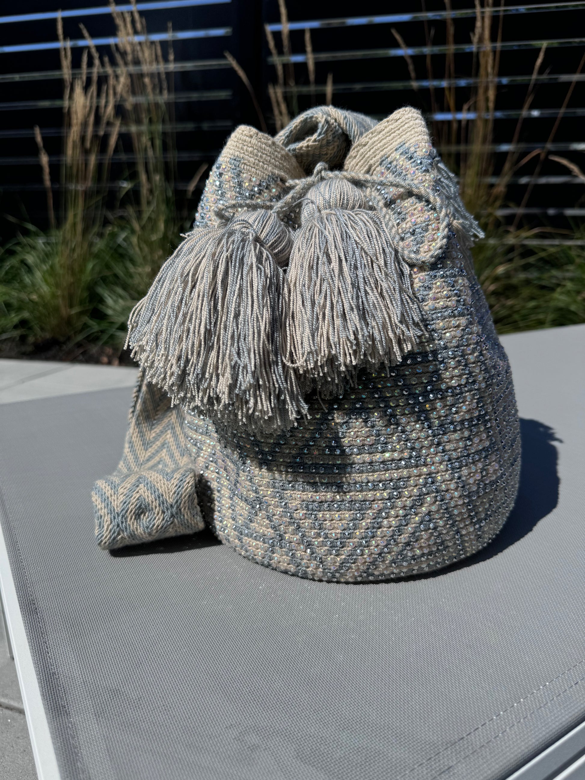 A light grey and beige geometric patterned shoulder bag with a fringed strap, placed on a flat surface with plants in the background.