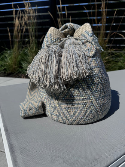 A light grey and beige geometric patterned shoulder bag with a fringed strap, placed on a flat surface with plants in the background.
