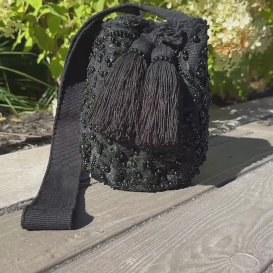 Black woven bag with tassel on a wooden surface with greenery in the background