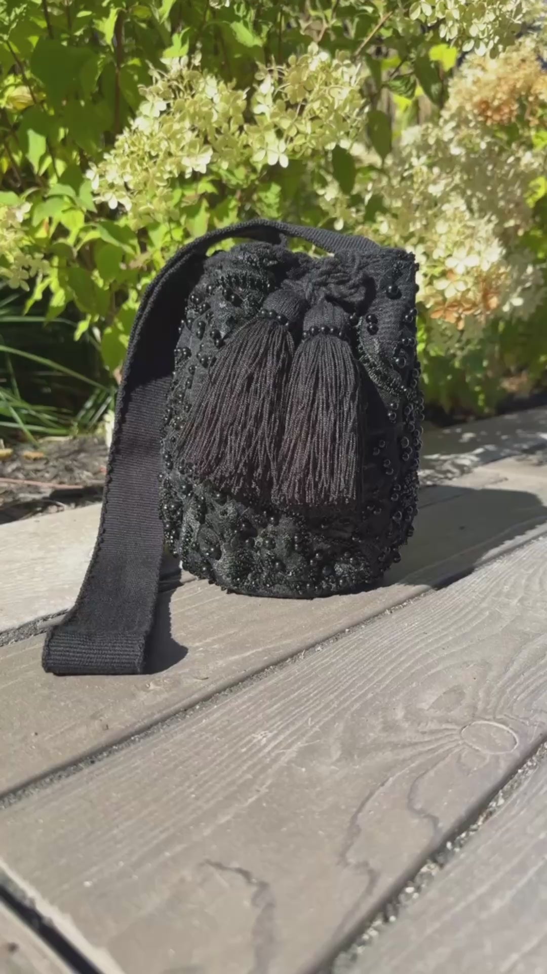 Black woven bag with tassel on a wooden surface with greenery in the background
