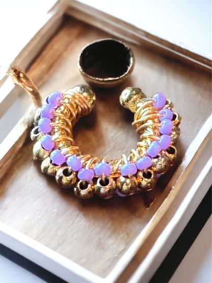 A purple and gold-colored beaded ear cuff displayed on a flat surface, with a small circular mirror reflecting part of the ear cuff.