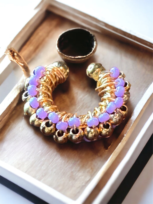 A purple and gold-colored beaded ear cuff displayed on a flat surface, with a small circular mirror reflecting part of the ear cuff.