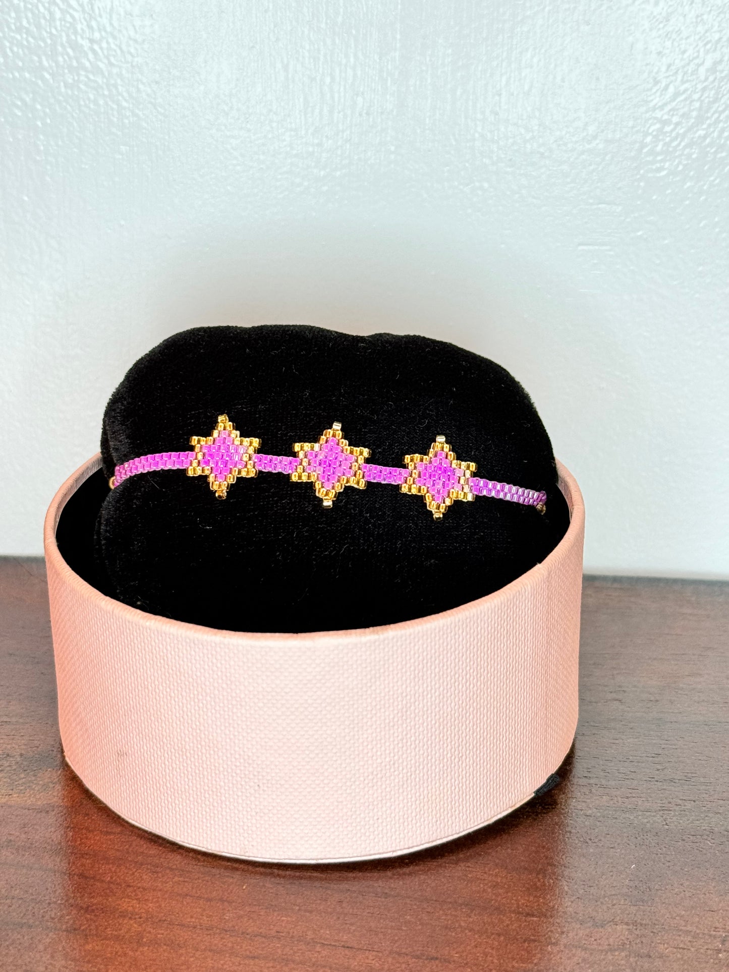 A bracelet with purple and gold colors displayed on a black cushion inside a pink box.