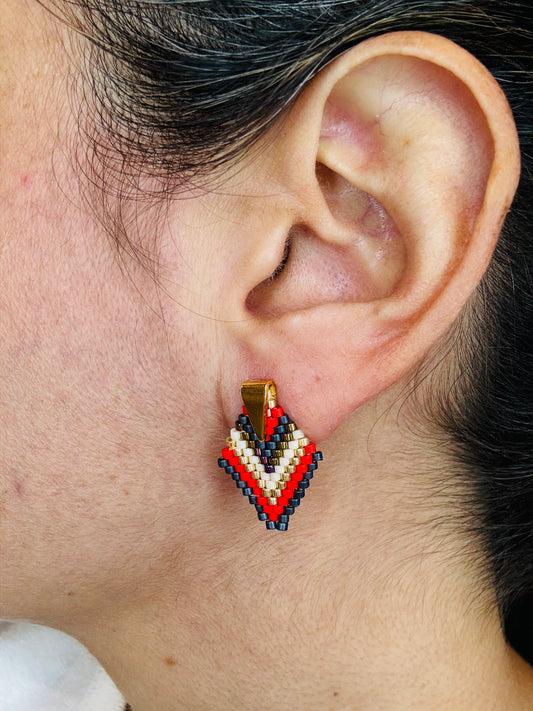 A person wearing a red, beige, and gold colored earring with a geometric pattern, made from crystal beads.