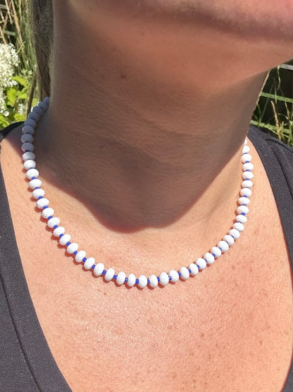 Close-up of a necklace with white and blue beads on a person's neck against a natural background.