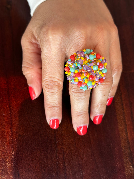 A colorful woven beaded ring displayed on a person's finger with red nail polish.