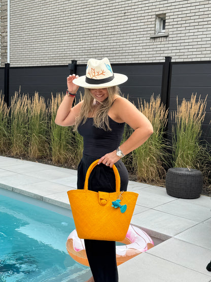 Woman by a pool holding a yellow bag and a white hat with a colorful design.